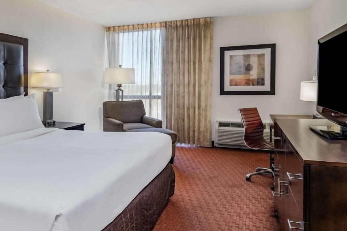 A hotel room with a large bed, a desk, a chair, a TV, and a window with curtains.