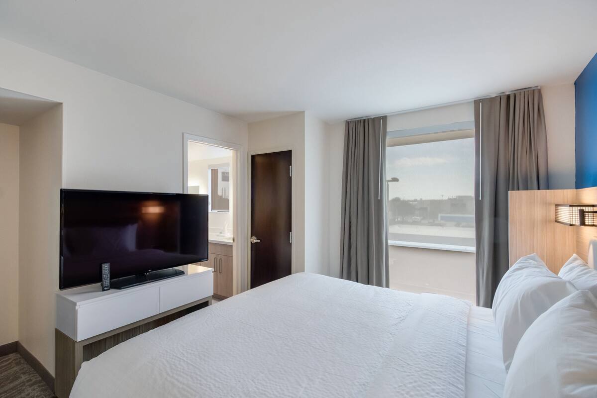 Habitación de hotel con una cama grande, un televisor de pantalla plana sobre una consola blanca, una puerta que da a un baño y una ventana con cortinas.
