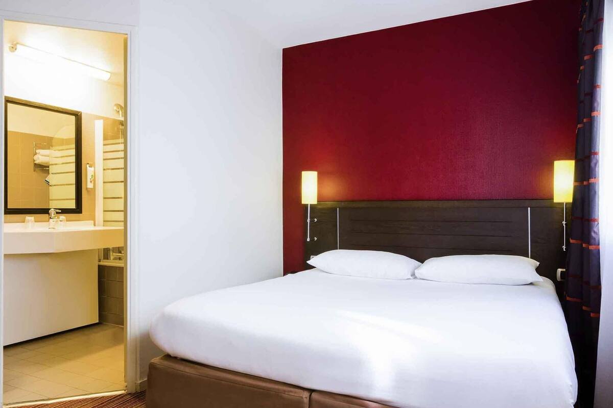 Une chambre d’hôtel avec un lit, une table de chevet, une lampe, un miroir et une salle de bain visible à travers une porte ouverte.