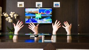 Une salle d’attente avec trois horloges au mur, un téléviseur diffusant une image colorée et un comptoir en verre derrière lequel se tiennent des personnes, les mains levées.