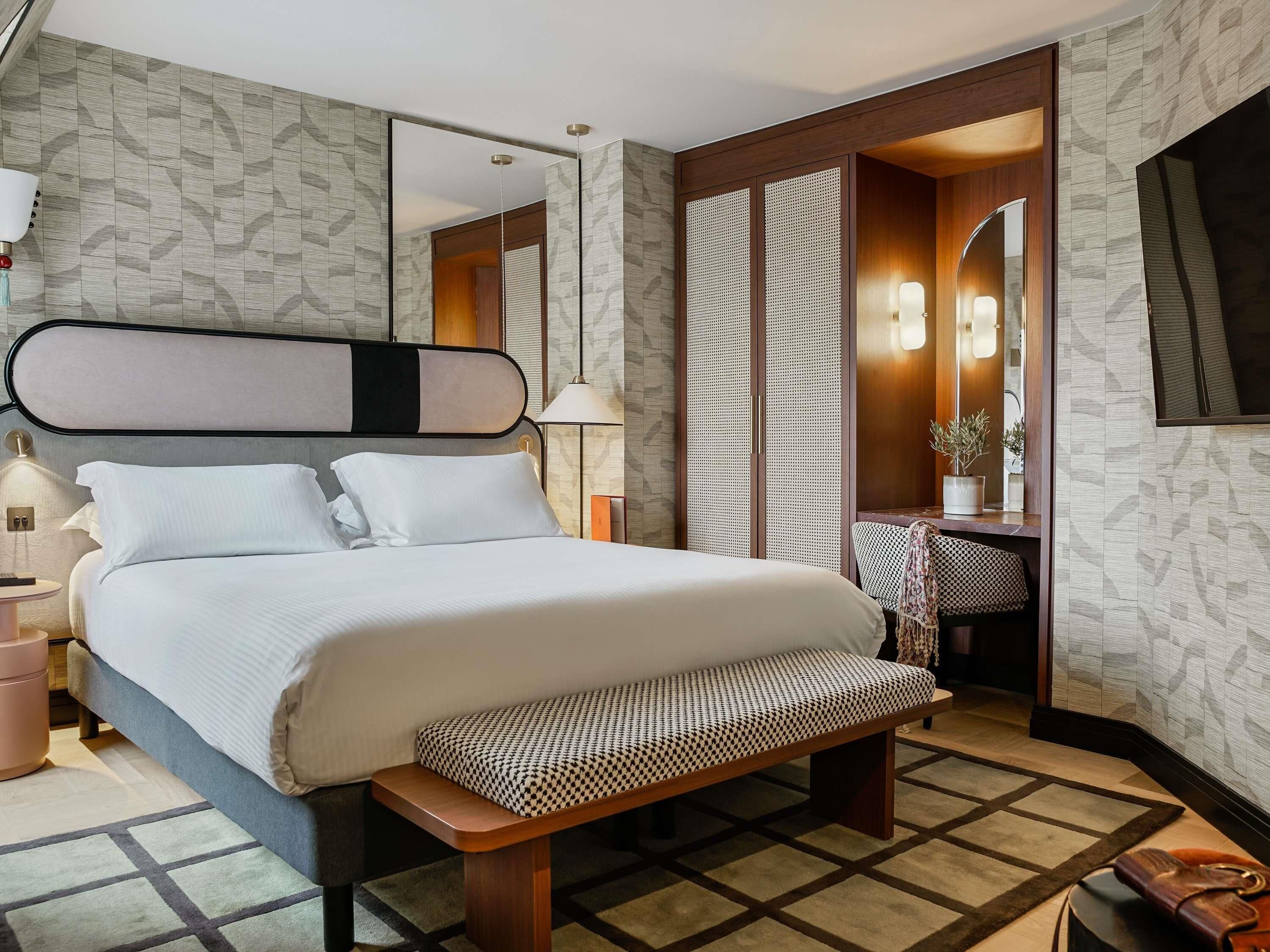 A hotel room with a large bed, bedside table, bench, and a mirror.