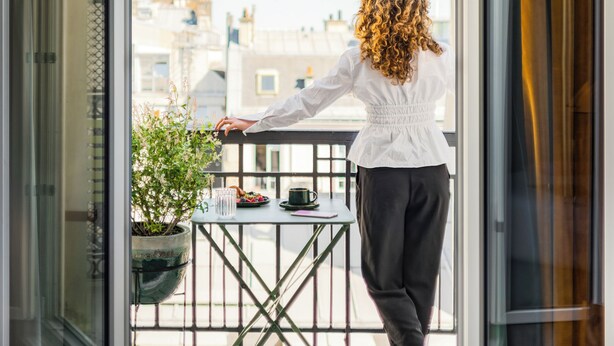 A person standing on a balcony with a city view, a small table with a plant, and a magazine on the bed. - Hotel Faubourg Galant Paris - Handwritten Collection (Paris)