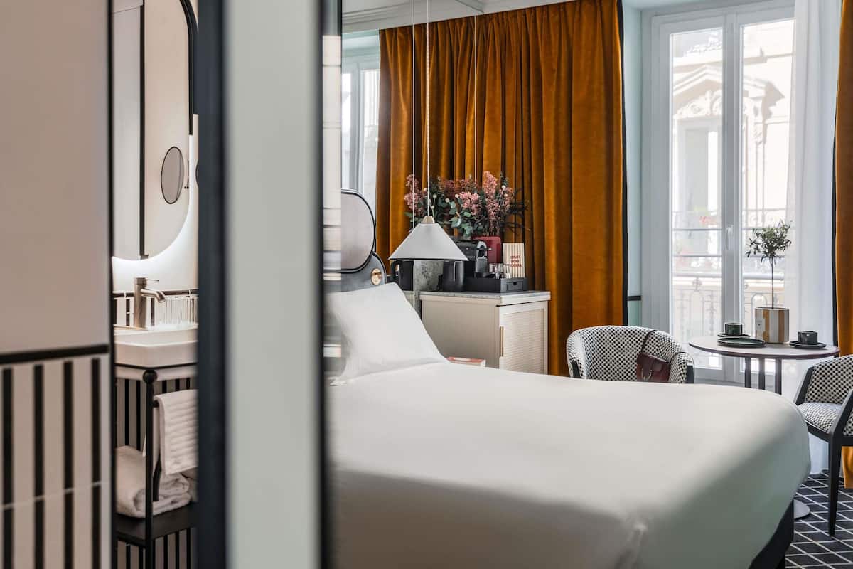 A hotel room with a bed, a desk, a chair, a small table, a lamp, and a window with curtains.