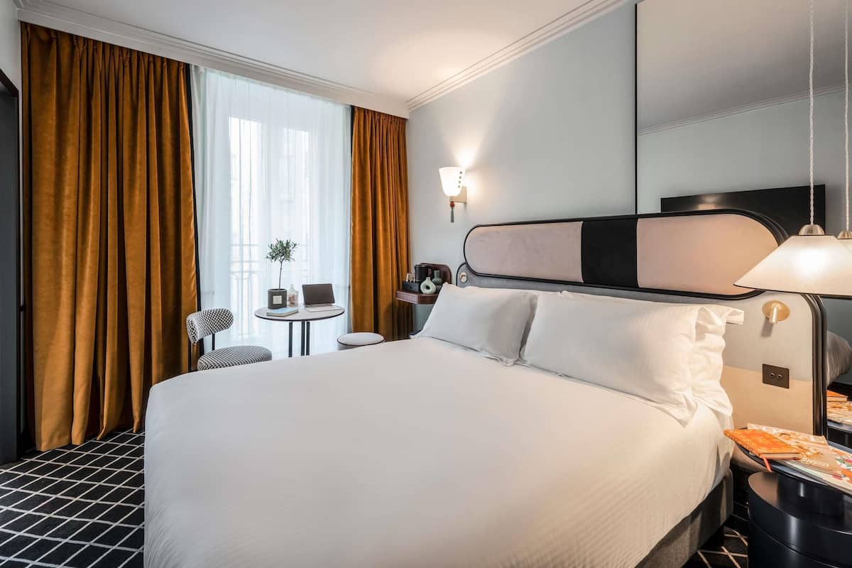 A hotel room with a large bed, a desk with a lamp, a chair, a small table with a plant, and a large mirror.