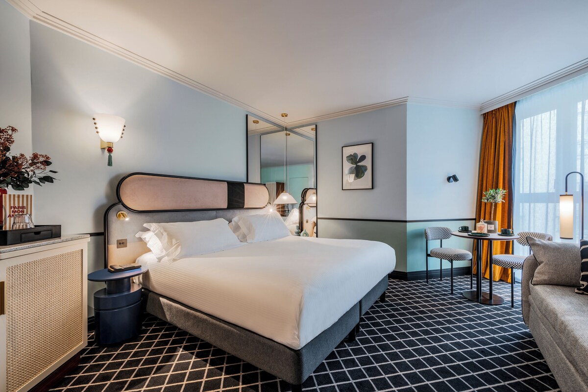 A hotel room with a large bed, bedside tables, a small round table with chairs, a mirror, and a window with curtains.