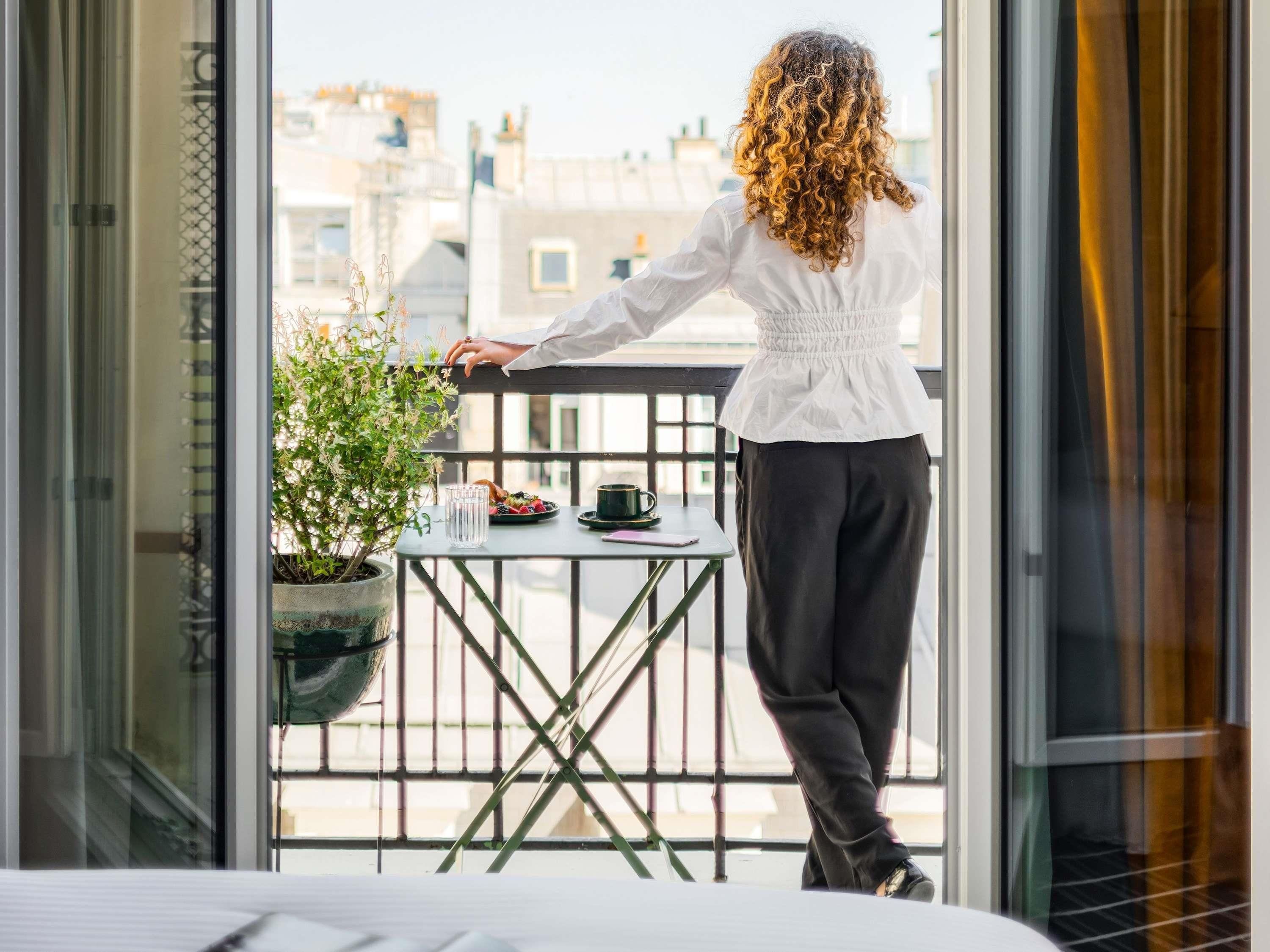 Une personne se tient sur un balcon, d’où l’on peut voir des bâtiments, une table avec une plante et une tasse.