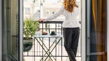 Une personne se tient sur un balcon, d’où l’on peut voir des bâtiments, une table avec une plante et une tasse.