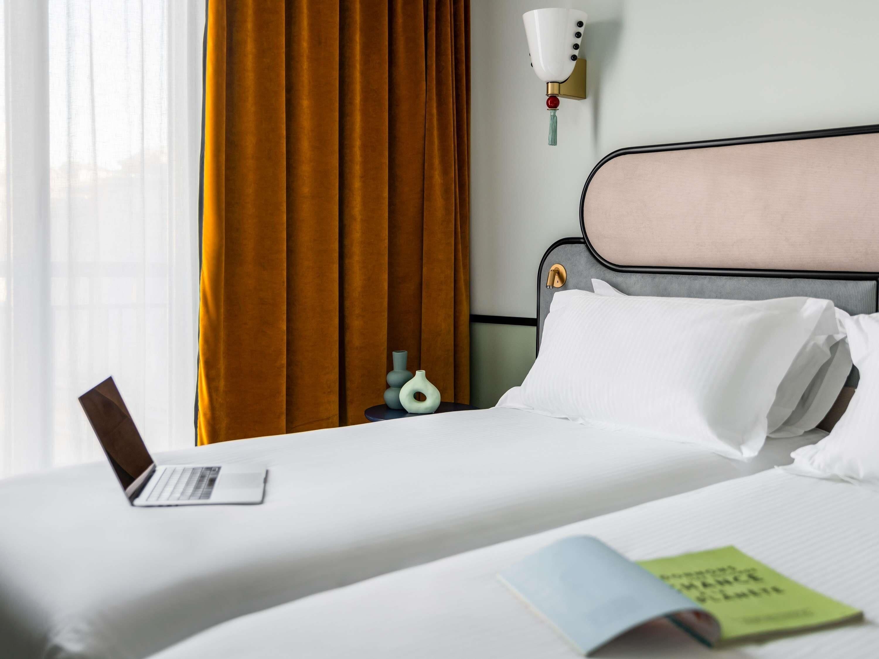 Una camera d'albergo con un letto, un portatile sul letto, un comodino con una lampada e una finestra con tende.