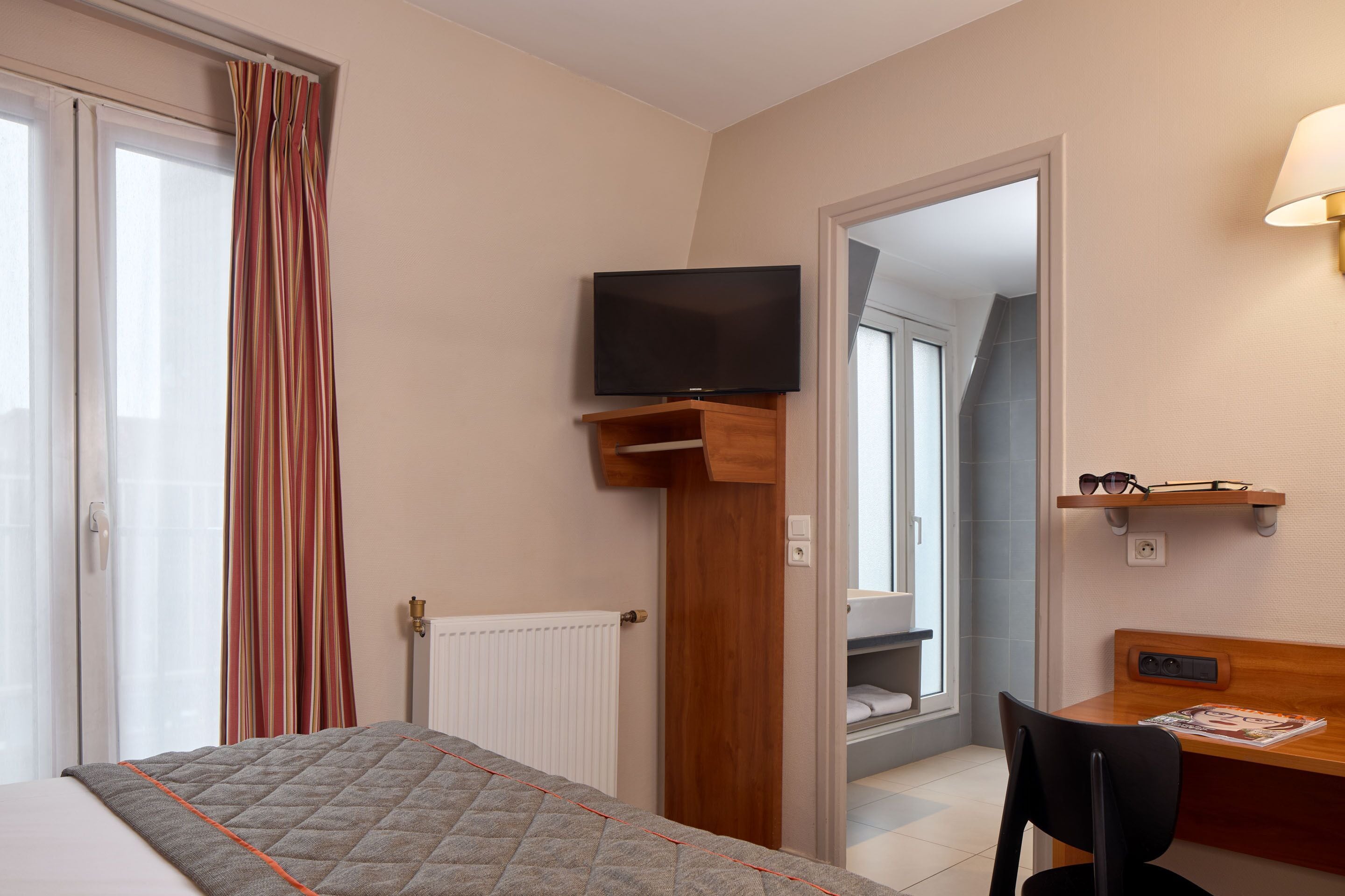 Habitación de hotel con cama, televisor montado en un estante de madera, escritorio con silla y espejo.