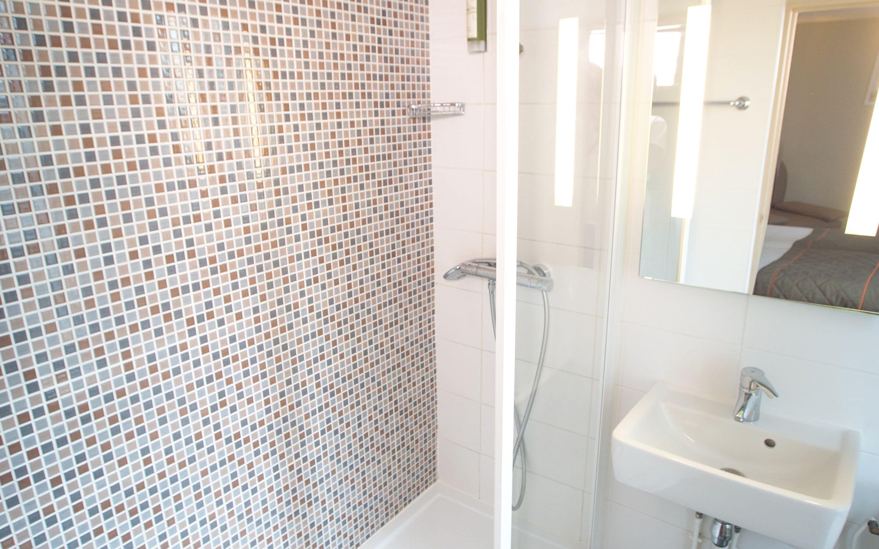 Une salle de bain avec un mur de douche recouvert de carreaux, une paroi de douche en verre et un lavabo blanc avec un miroir au-dessus.