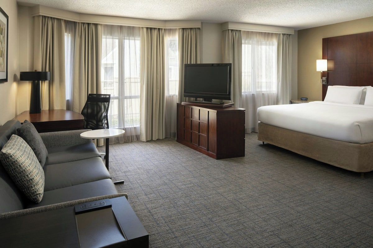 A hotel room with a bed, a sofa, a TV on a wooden cabinet, a desk with a lamp, and a window with curtains.