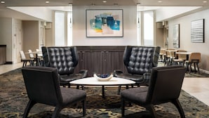 Un hall d’hôtel moderne avec une table ronde, deux fauteuils en cuir, un saladier de fruits et un grand tableau abstrait accroché au mur.