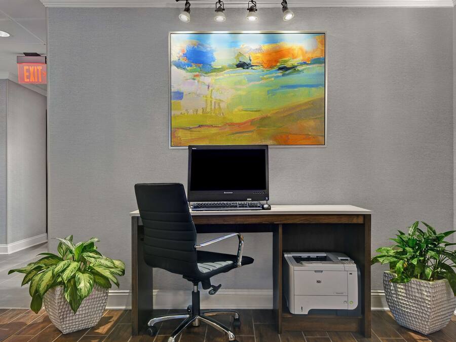 A modern office space with a desktop computer, a black office chair, two potted plants, and a large abstract painting on the wall.