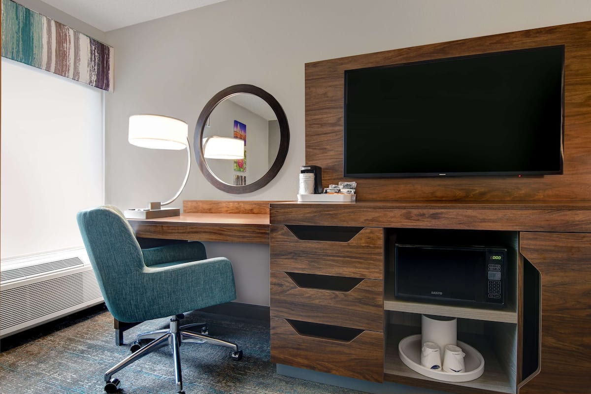 A hotel room with a teal chair, a desk with a lamp, a flat-screen TV, a microwave, and a coffee maker.