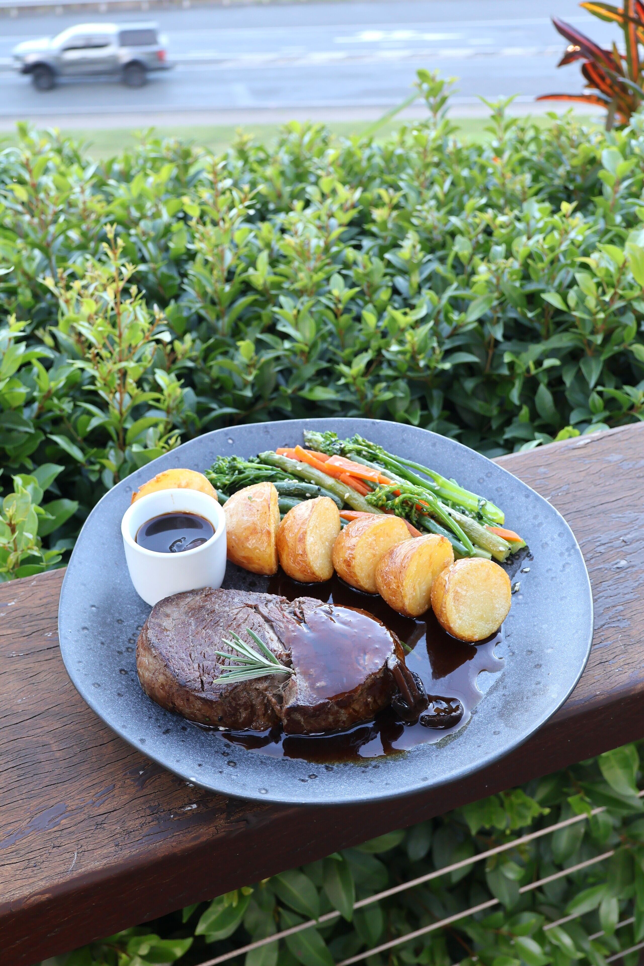 A plate of food with steak, vegetables, and sauce on a table with a view of a road and greenery.