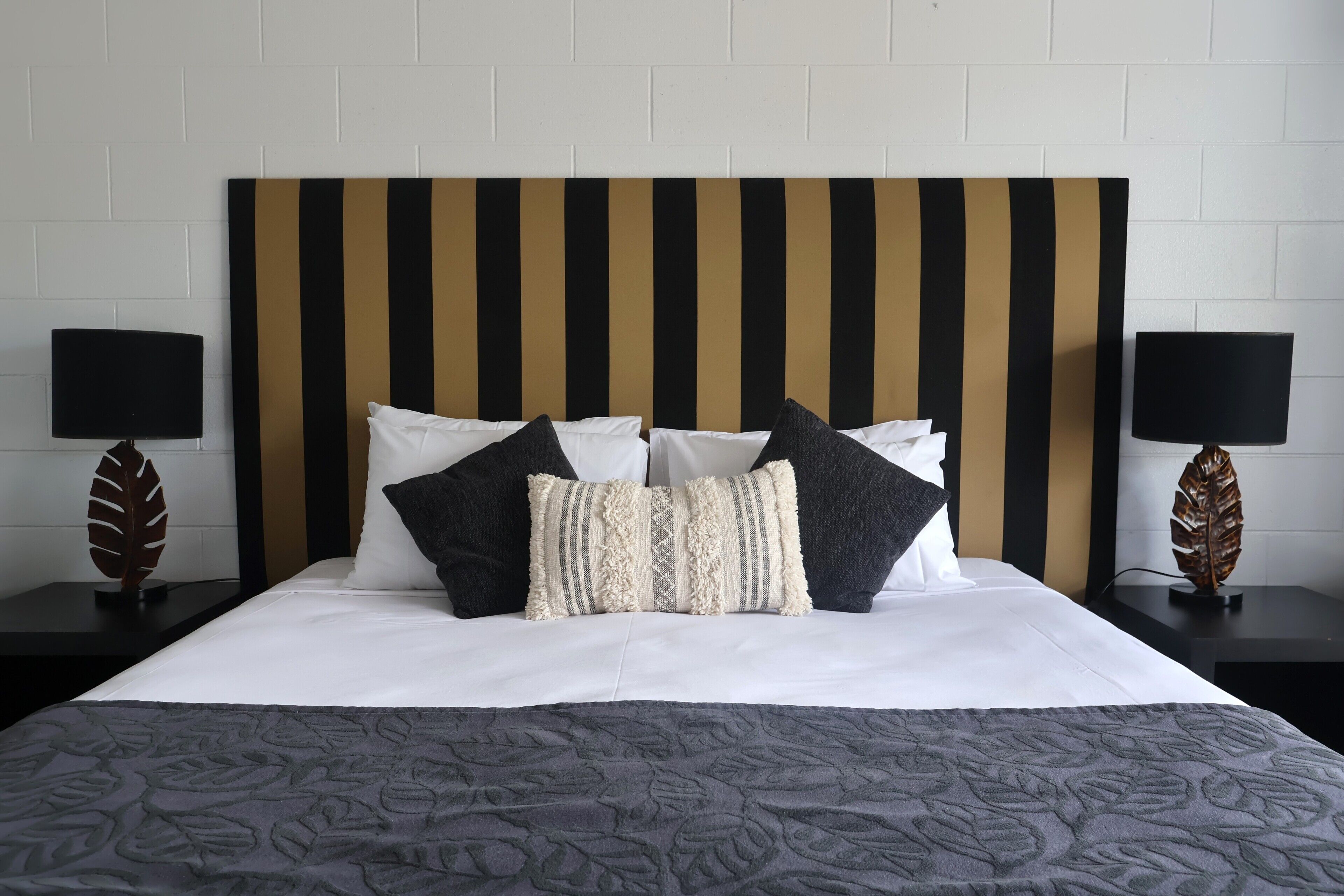 Una cama bien hecha con un cabecero a rayas, dos mesitas de noche con lámparas y objetos decorativos con forma de hoja sobre las mesitas.