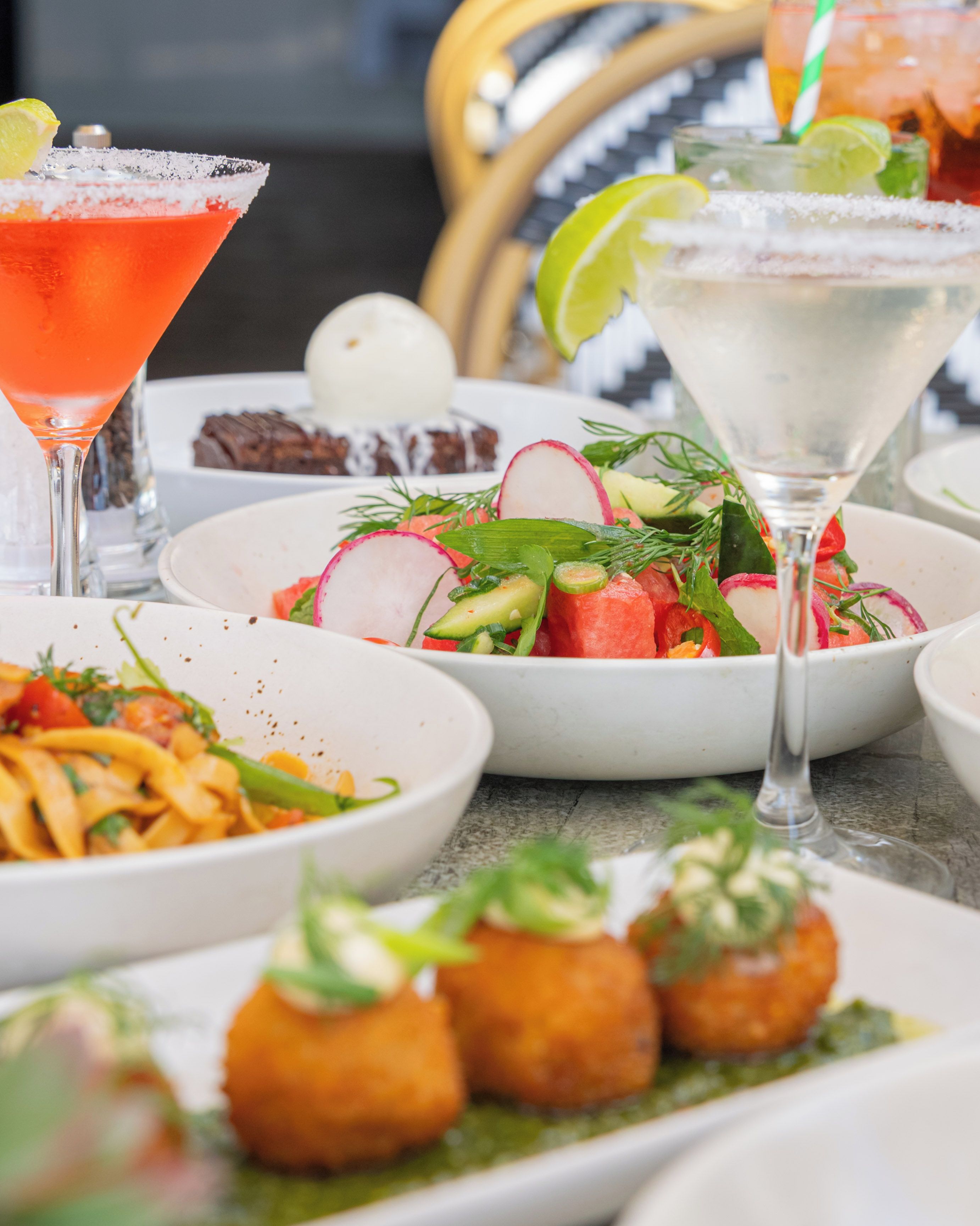 Une table garnie de divers plats, notamment une salade, des pâtes et des amuse-gueule, ainsi que deux cocktails.