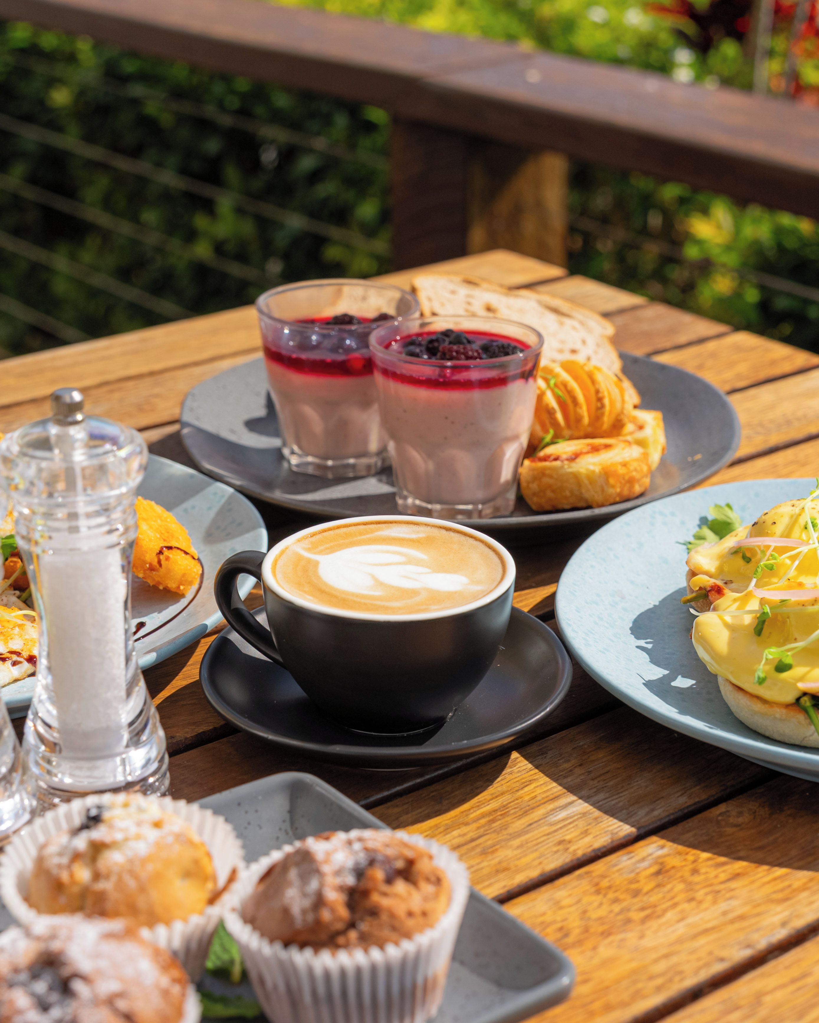 A wooden table with a variety of food items including pastries, a cup of coffee, and a glass of a red beverage.