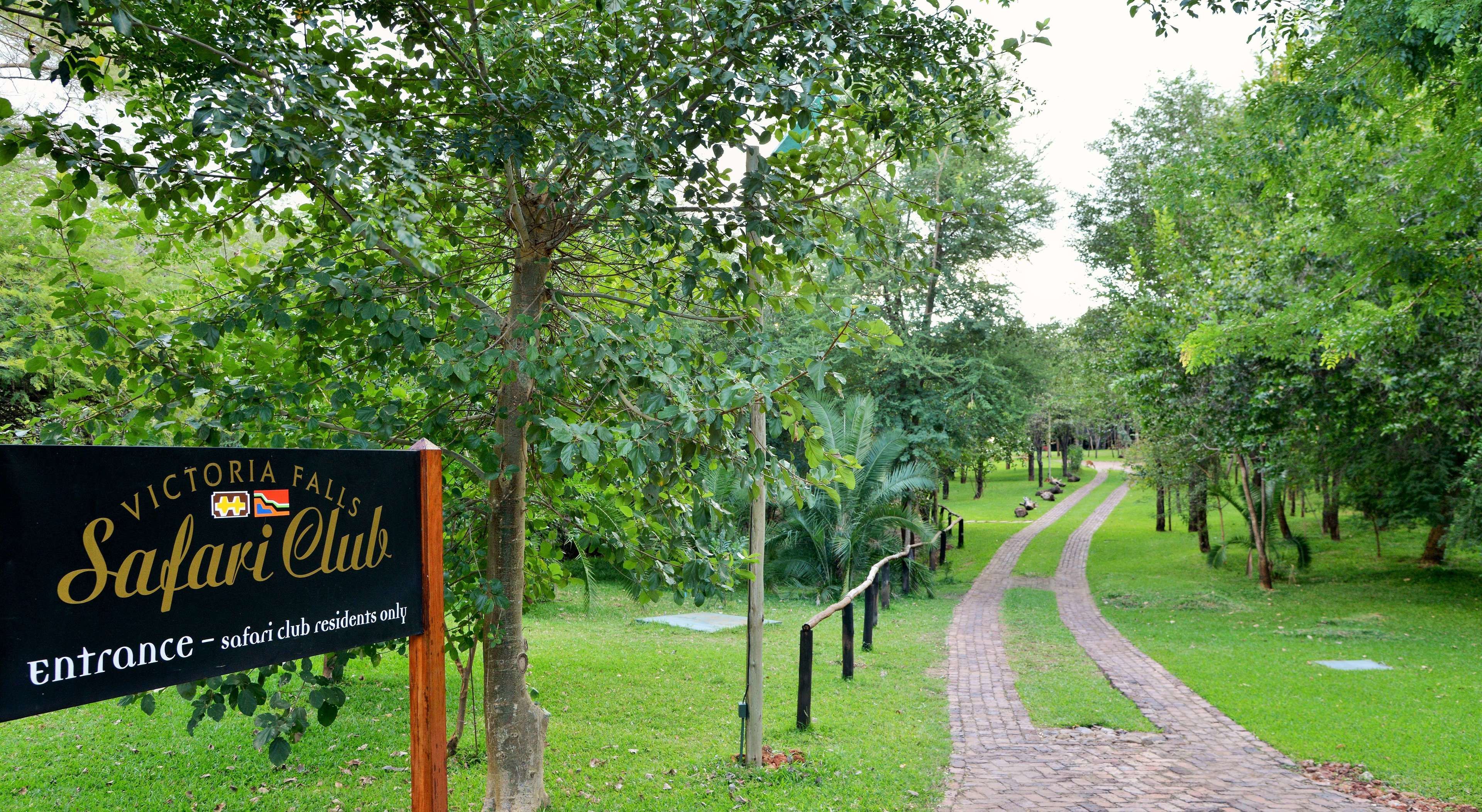 A sign for Victoria Falls Safari Club with a brick pathway leading through a lush green area.