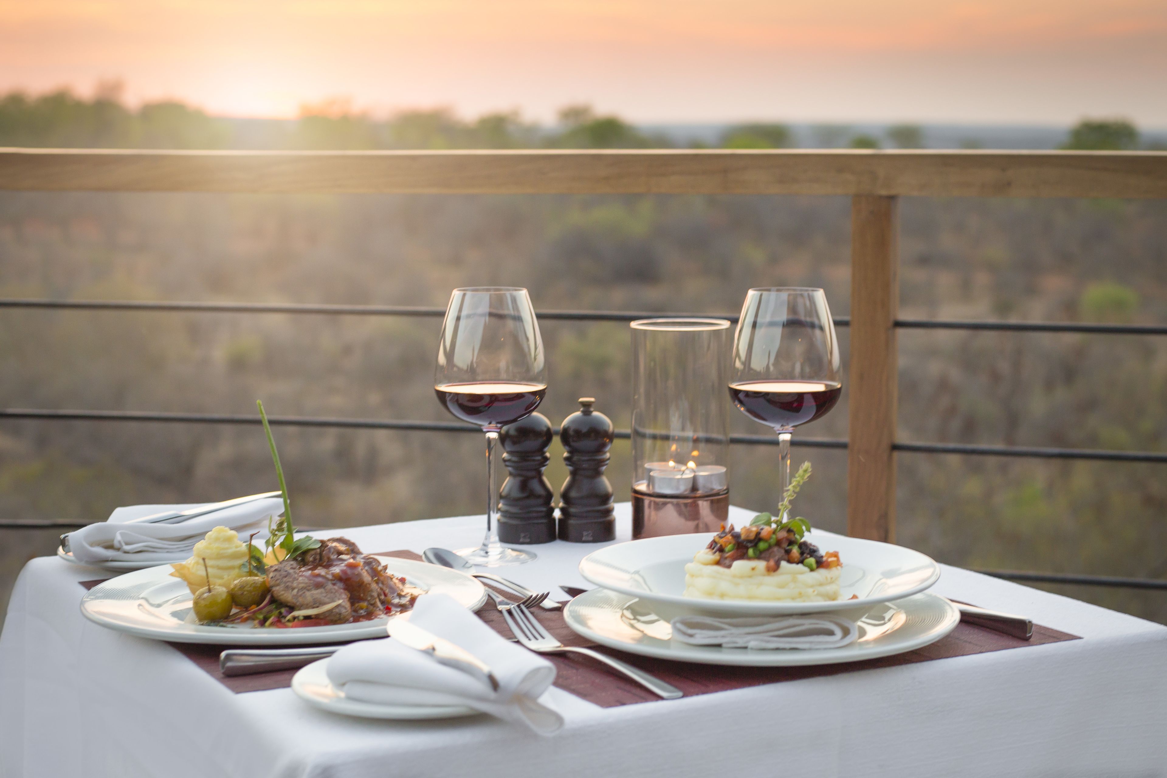A table set for a meal with two plates of food, wine glasses, and salt and pepper shakers on a balcony with a scenic view.