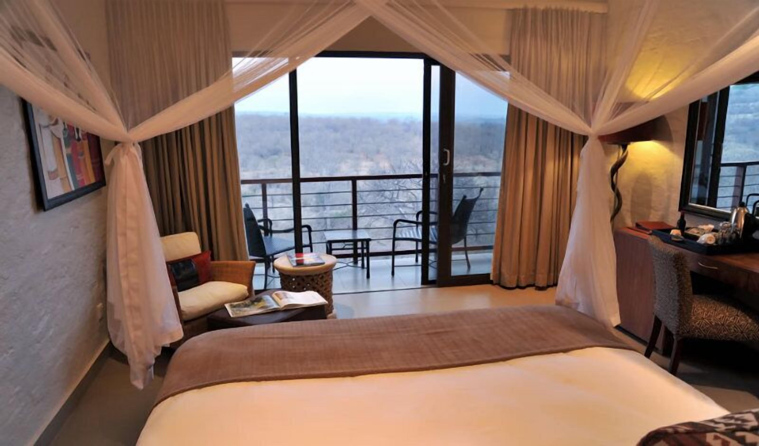 A hotel room with a large bed, a small table with a magazine, a chair, and a view of the outdoors through large windows.