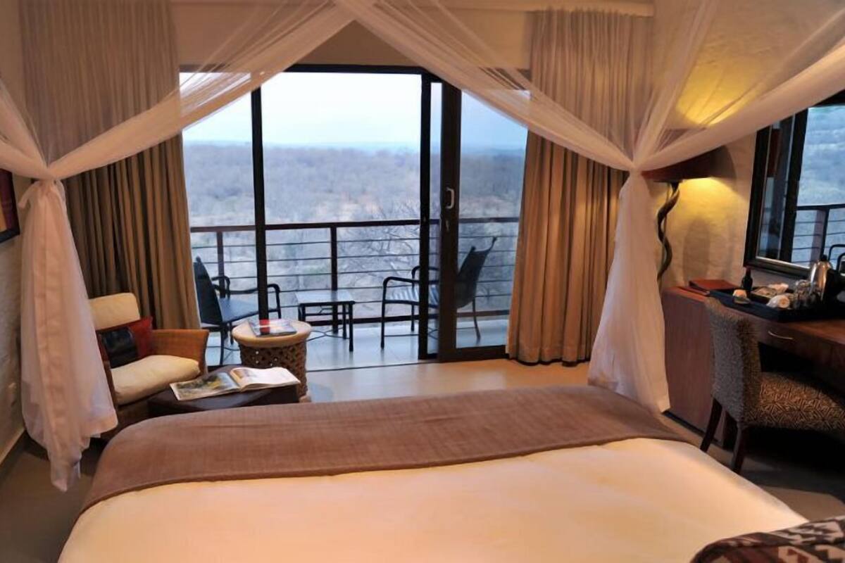 A hotel room with a large bed, a small table, a chair, and a balcony with a view.