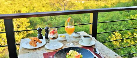 A breakfast table with croissants, a fruit parfait, a glass of orange juice, and a plate of eggs Benedict, set against a scenic view of a forested landscape.