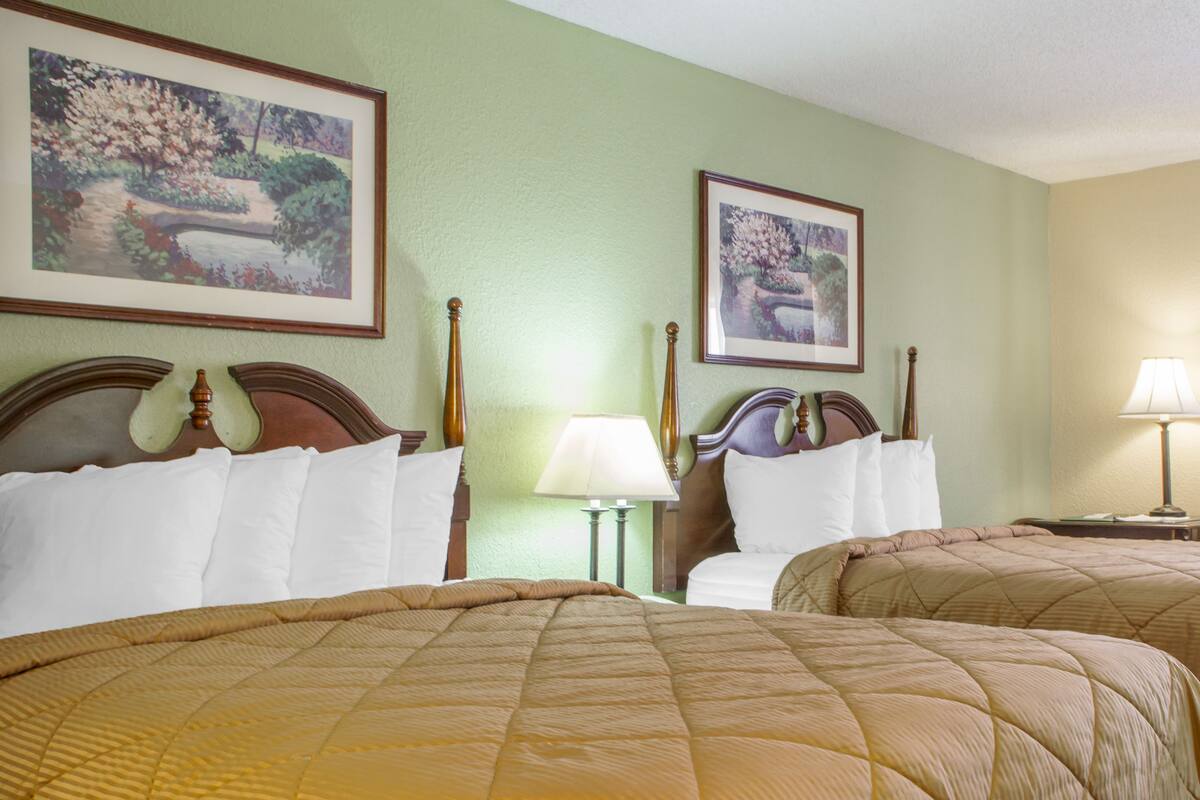 Quarto de hotel com duas camas de casal, uma mesinha de cabeceira com abajur e duas pinturas emolduradas na parede.
