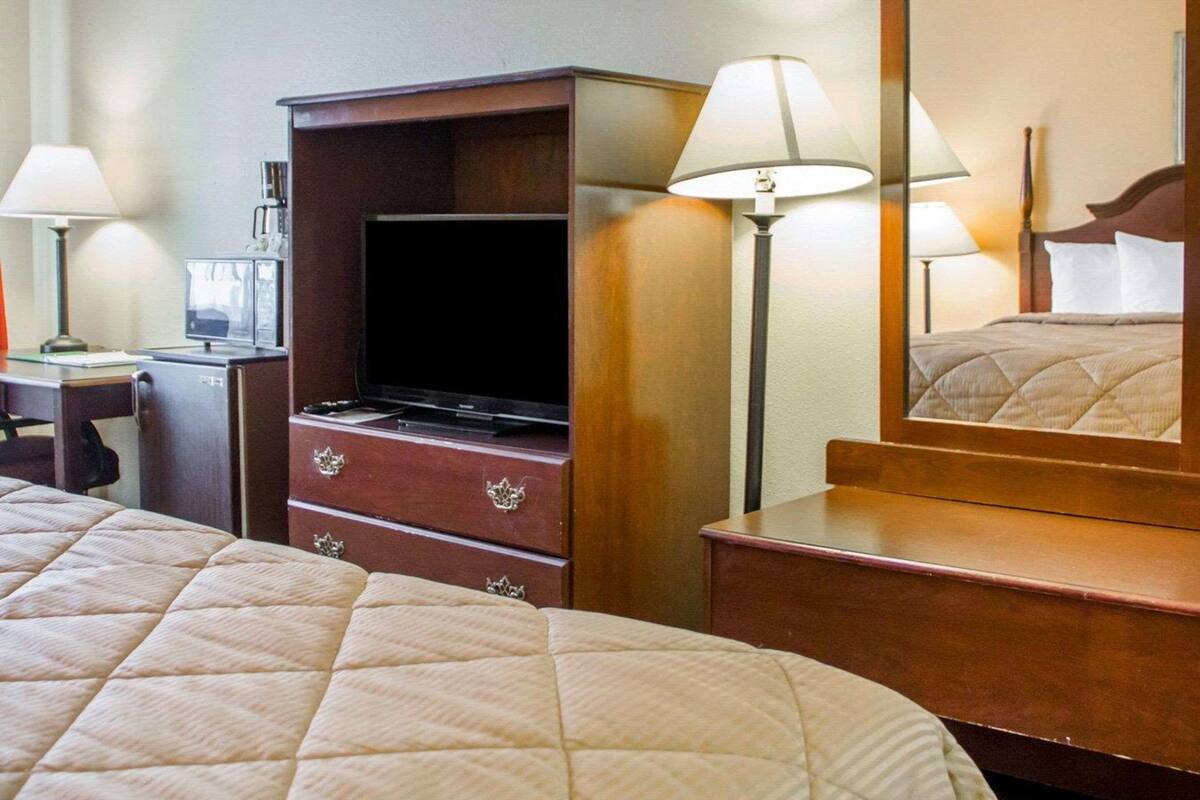 Quarto de hotel com cama, escrivaninha com computador, televisão, cômoda e espelho.