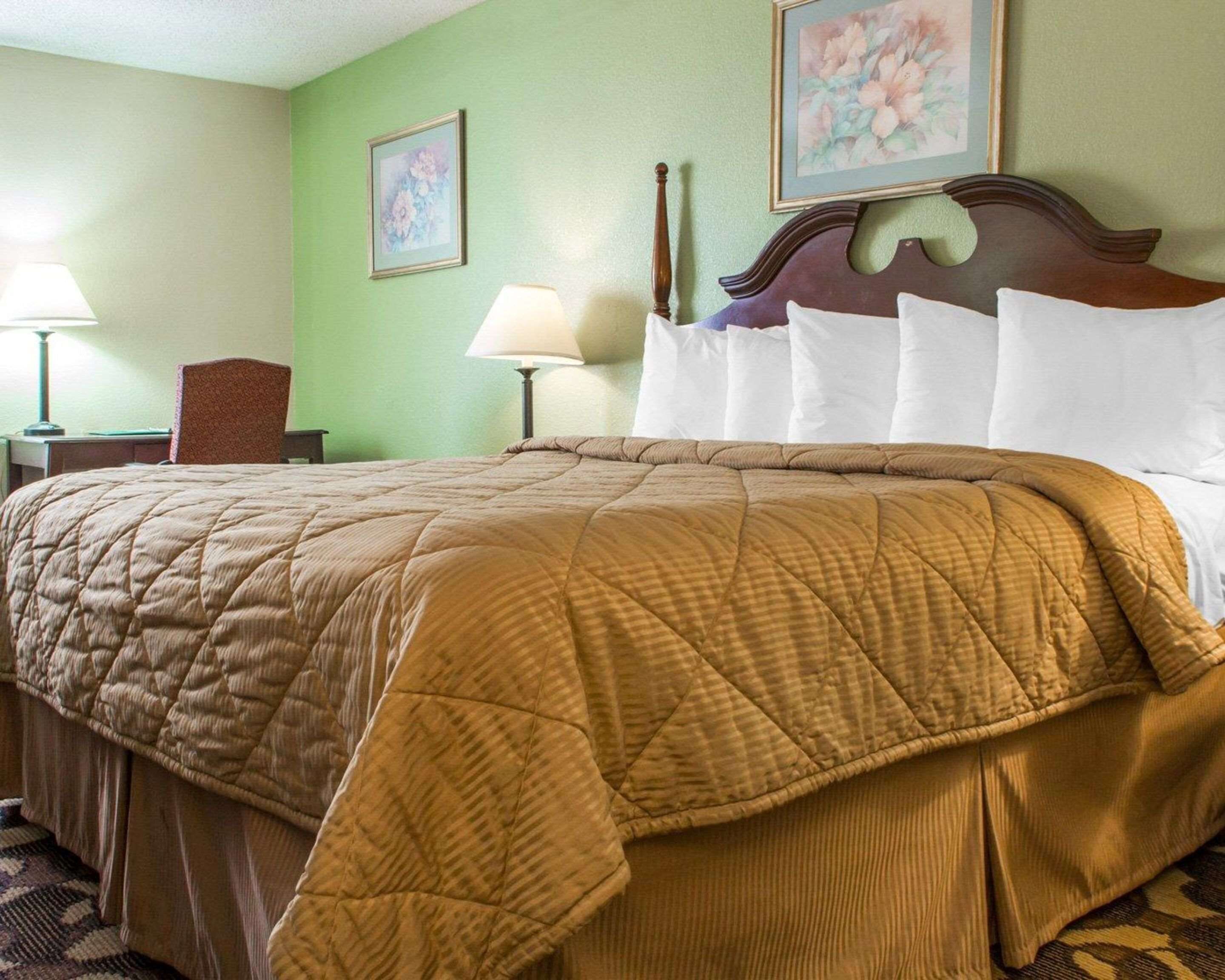 A neatly made bed with a brown quilt, a wooden headboard, and white pillows. There are two wall-mounted lamps and a framed floral painting above the bed.