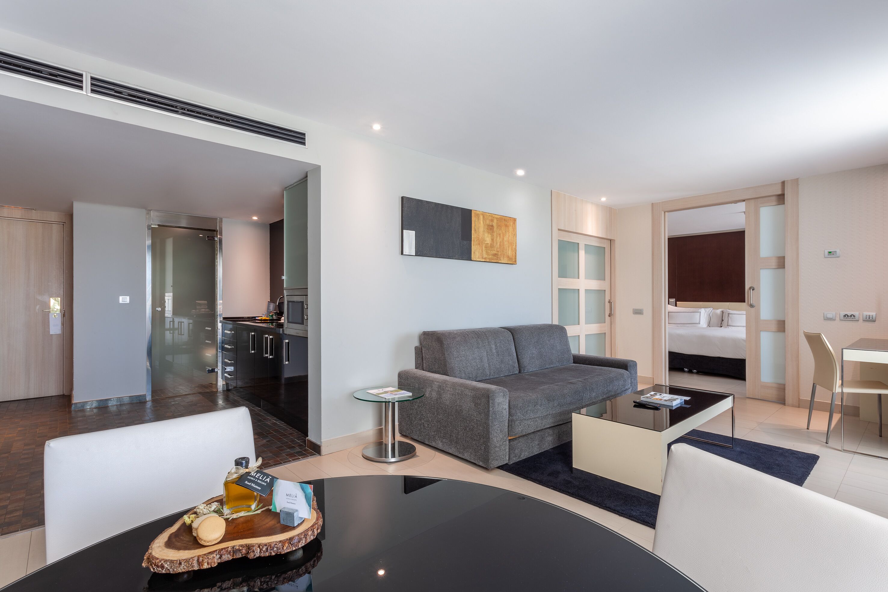 Una habitación de hotel moderna con un sofá gris, una pequeña mesa redonda con una bandeja de aperitivos, un televisor de pantalla plana montado en la pared y un área de comedor separada con una silla y una mesa.