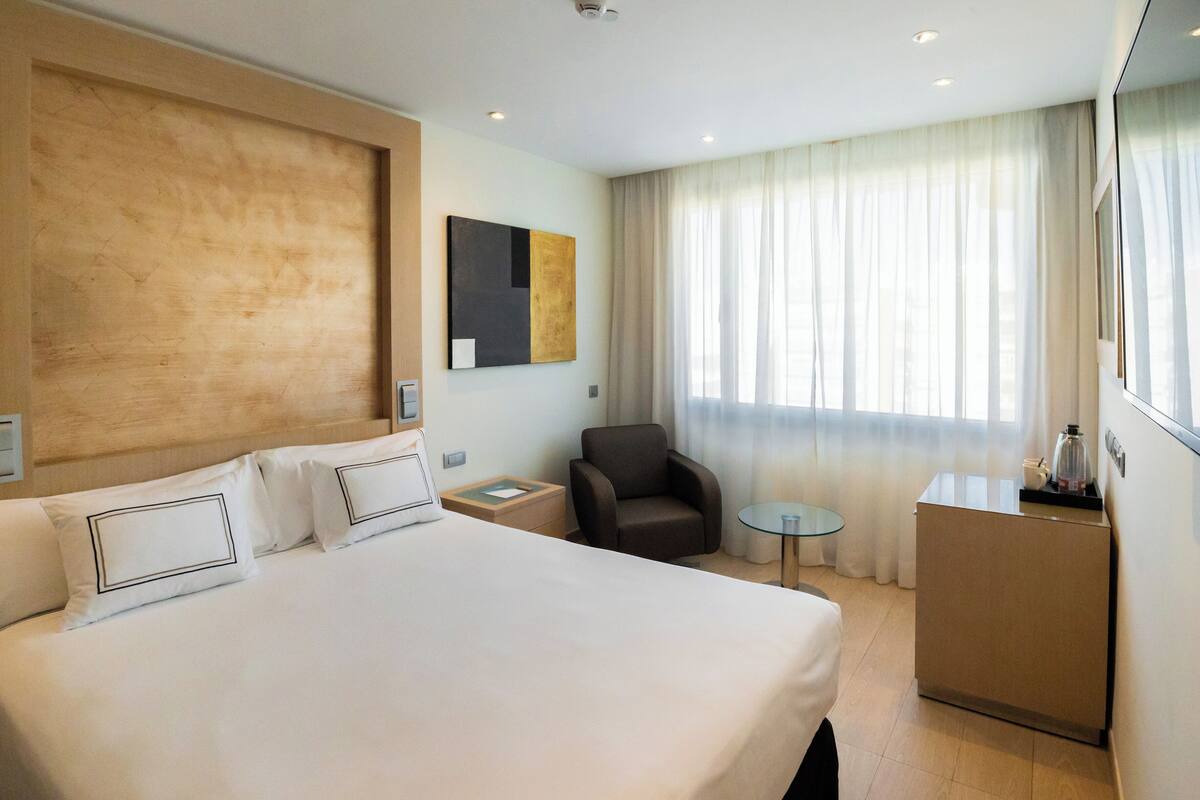 A hotel room with a large bed, a small desk, a chair, and a window with curtains.