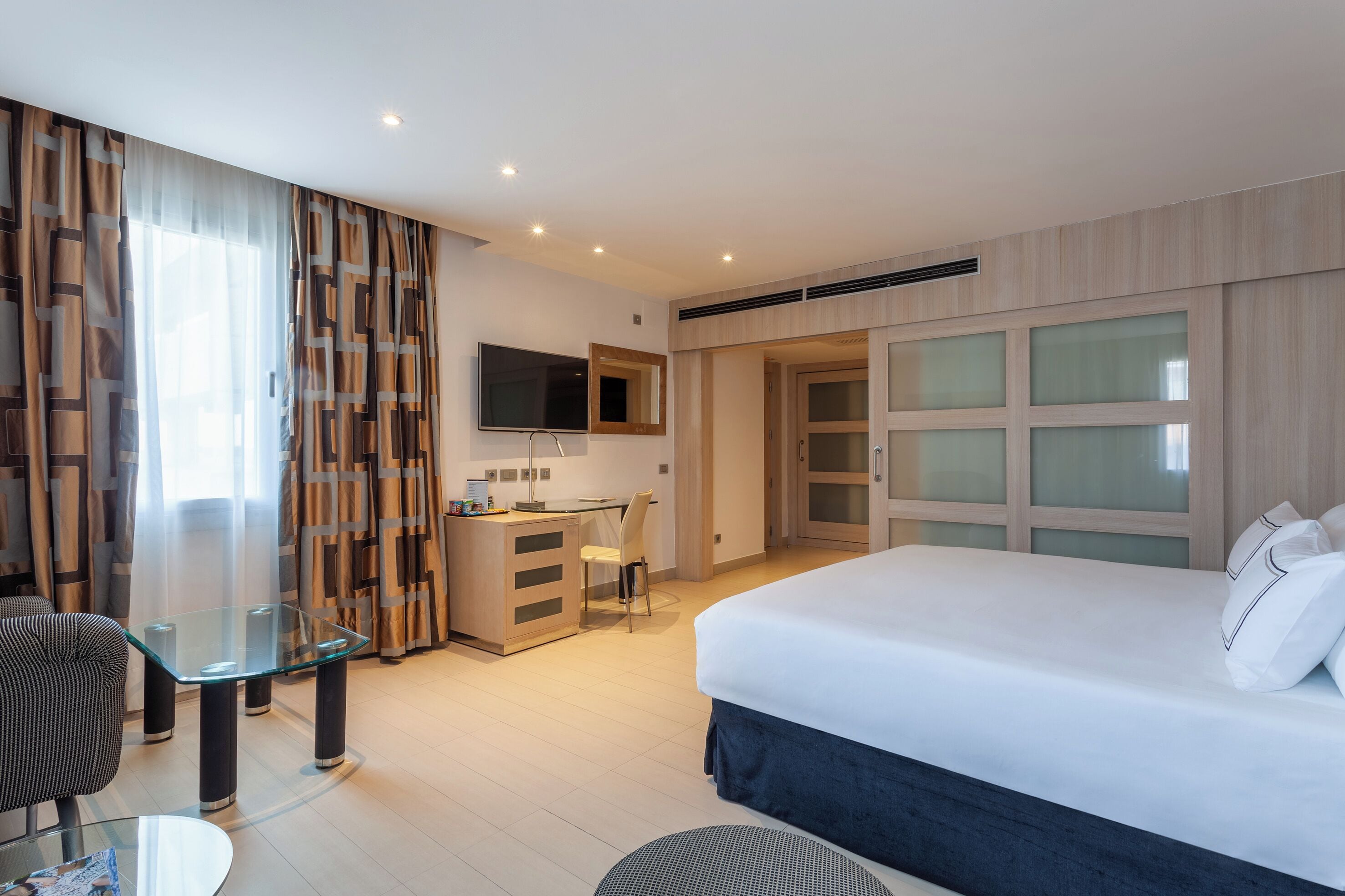 Una habitación de hotel moderna con una cama grande, un escritorio con una silla, un televisor y un armario con puertas de vidrio.