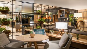 A modern living room with a round wooden table, grey upholstered chairs, and a sofa. There are multiple hanging plants and a large whiteboard on a stand. - Melia Sevilla (Seville)