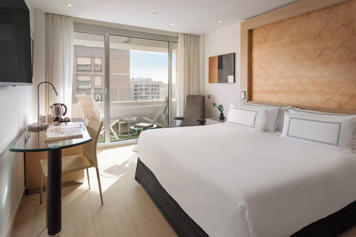 A hotel room with a large bed, a glass desk, a chair, a TV, and a view of the city through large windows.