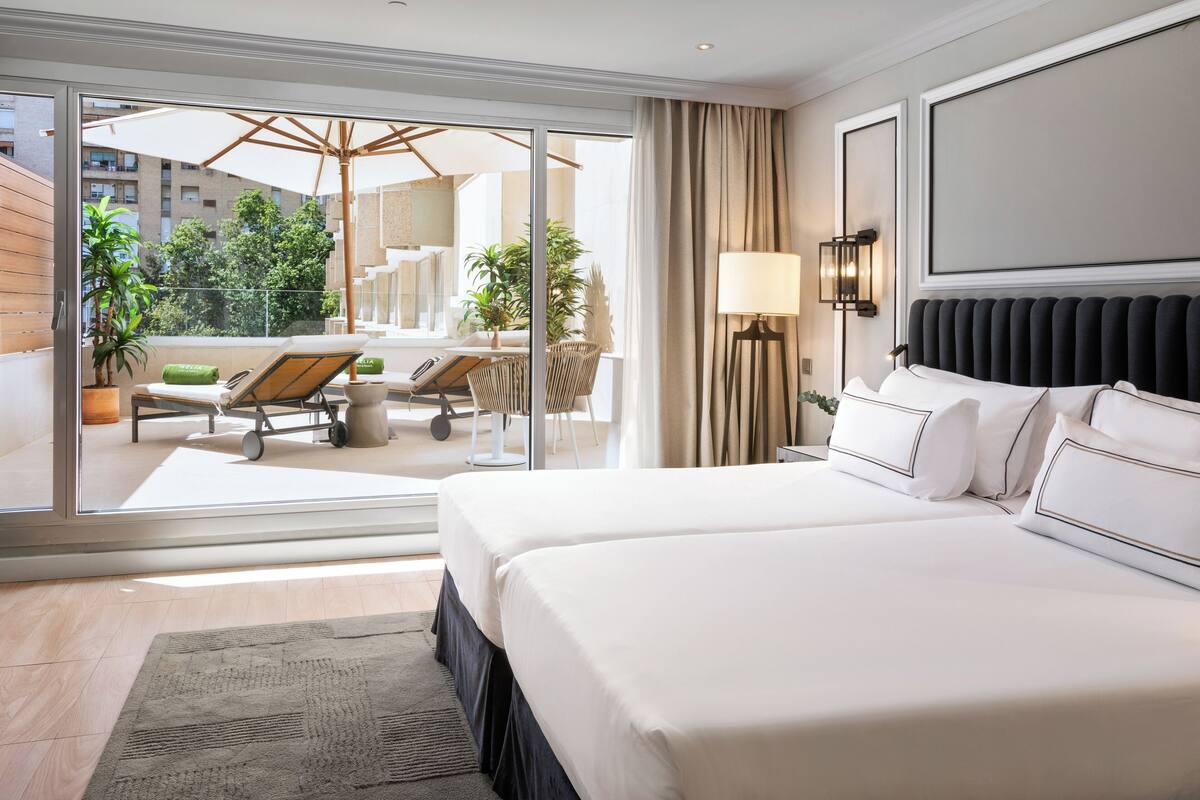 A hotel room with a large bed, a balcony with a sun lounger, and a view of the cityscape.