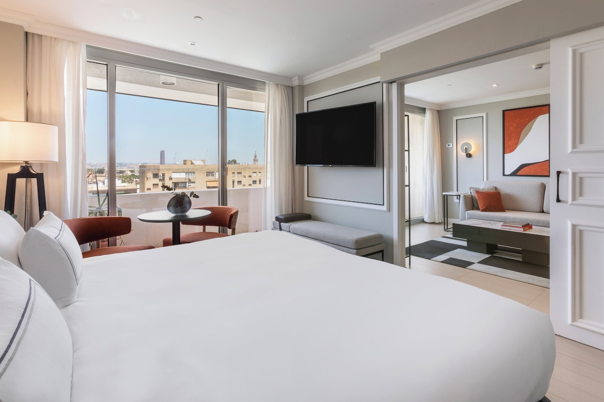 A hotel room with a large bed, a desk with a chair, a sofa, a TV mounted on the wall, and a view of the city through large windows.