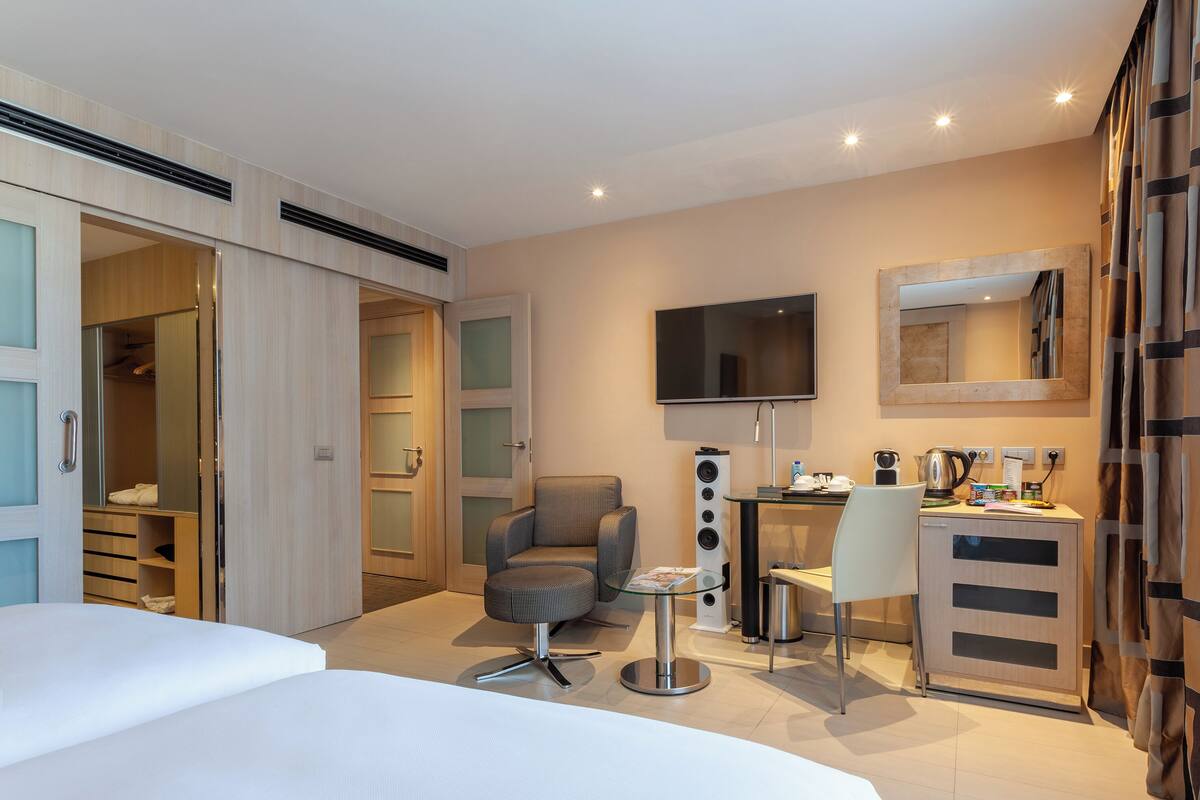 A modern hotel room with a bed, a desk with a computer, a chair, a TV, and a wardrobe.