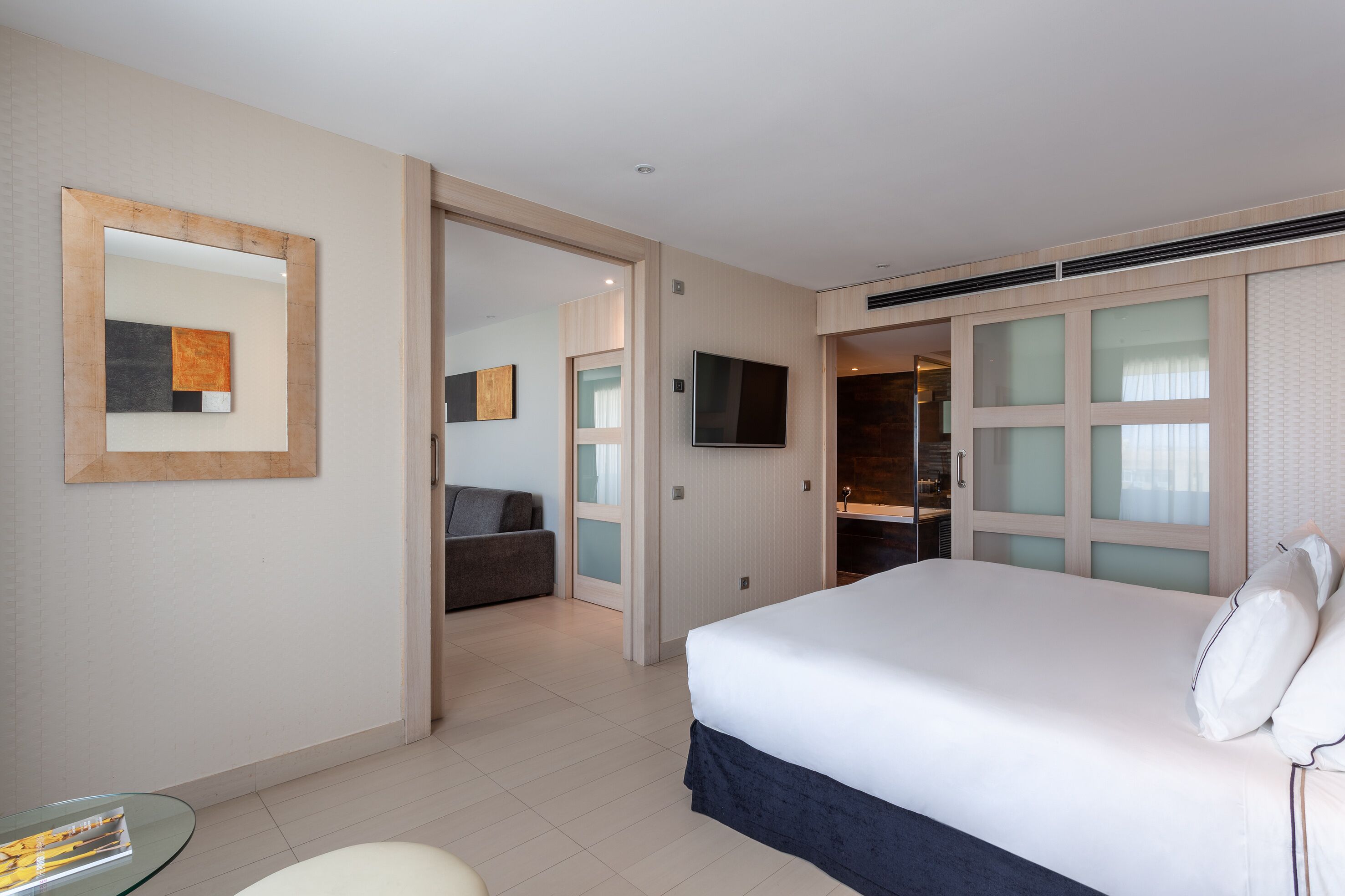 A hotel room with a large bed, a TV mounted on the wall, a glass table, and a seating area with a couch.
