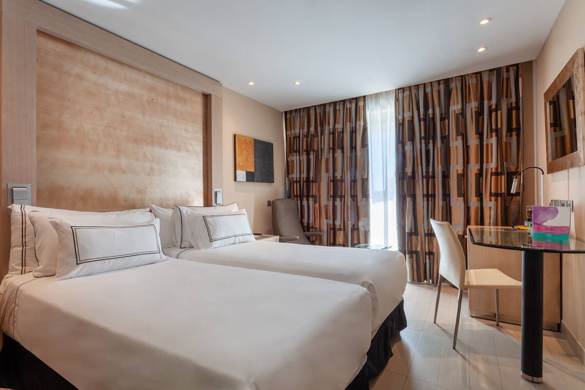 A hotel room with two beds, a desk with a chair, a large window with curtains, and a painting on the wall.