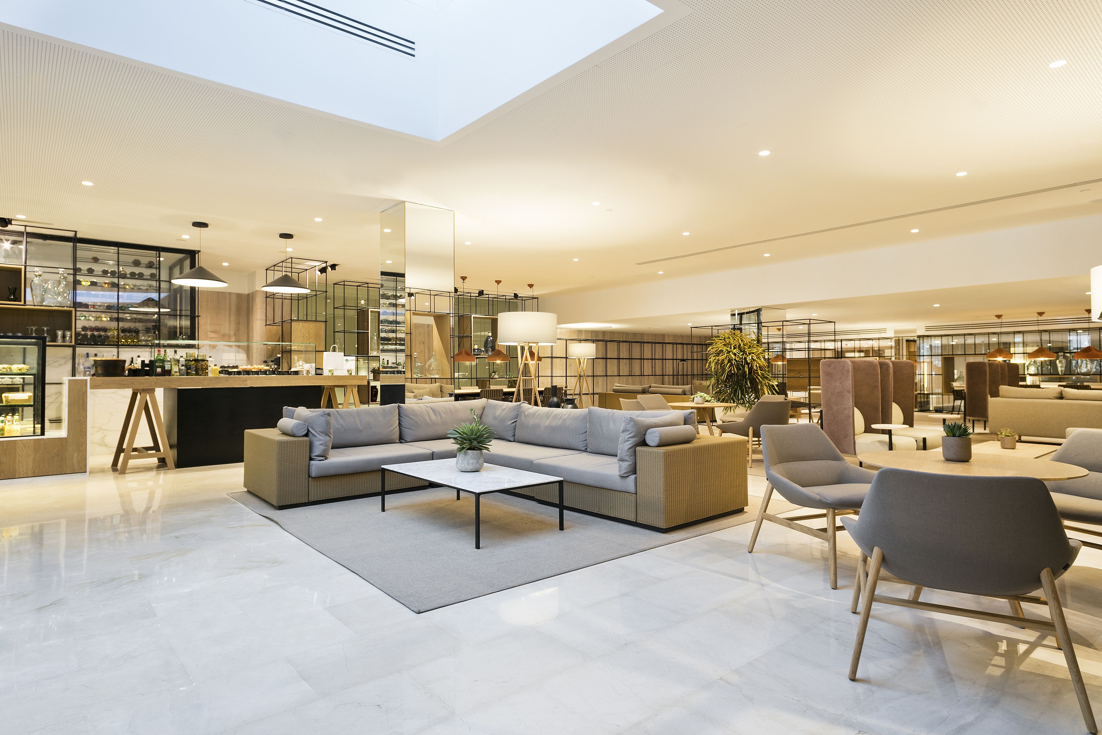 Un'ampia hall d'hotel moderna, con soffitto alto, zona salotto centrale con divani e poltrone, area bar e un grande specchio che riflette l'ambiente.