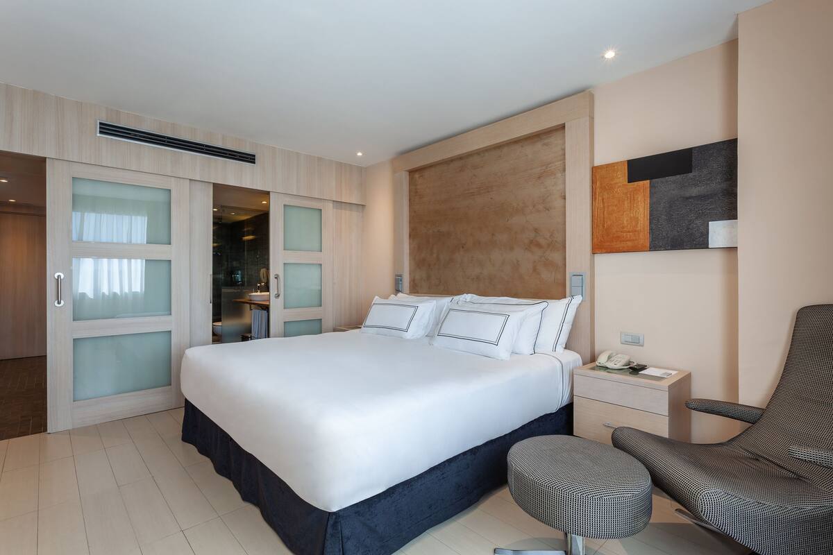 A modern hotel room with a large bed, a desk, a chair, and a wardrobe.