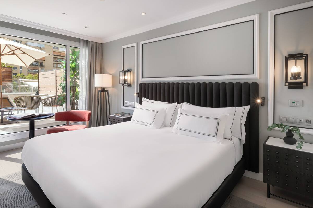 A modern hotel room with a large bed, a small desk, a chair, and a view of the outside.