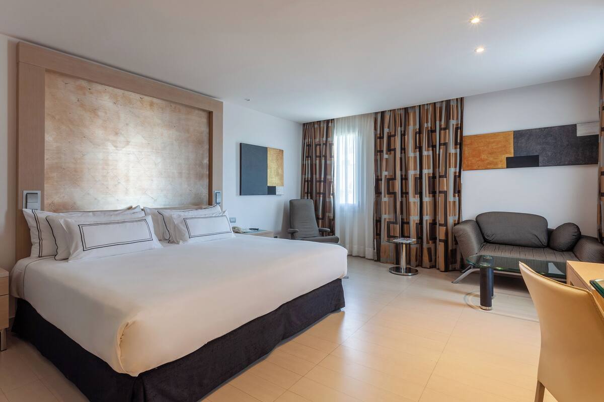 A modern hotel room with a large bed, a sofa, a glass coffee table, and a TV.