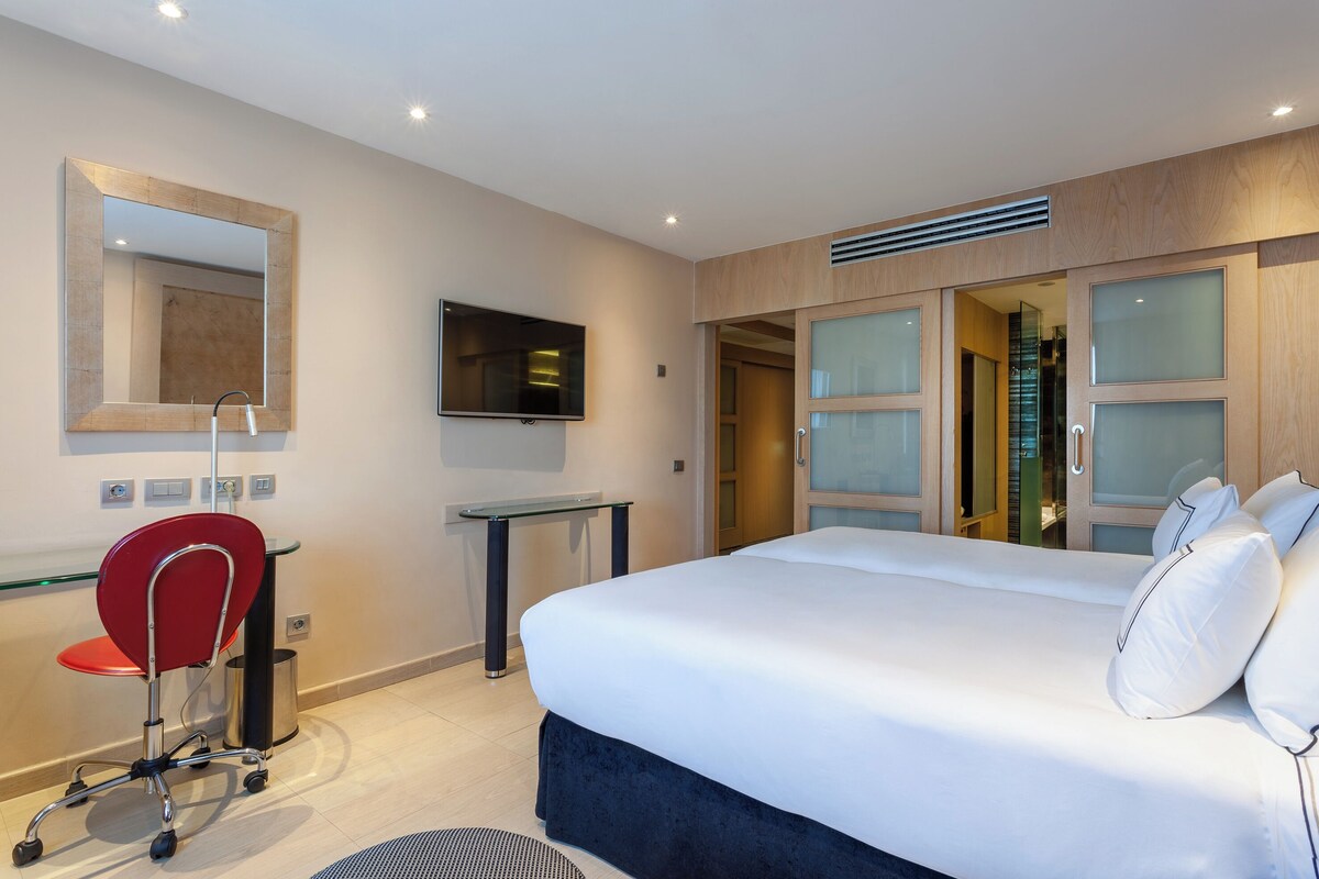 A modern hotel room with a large bed, a desk with a chair, a TV, and a mirror.