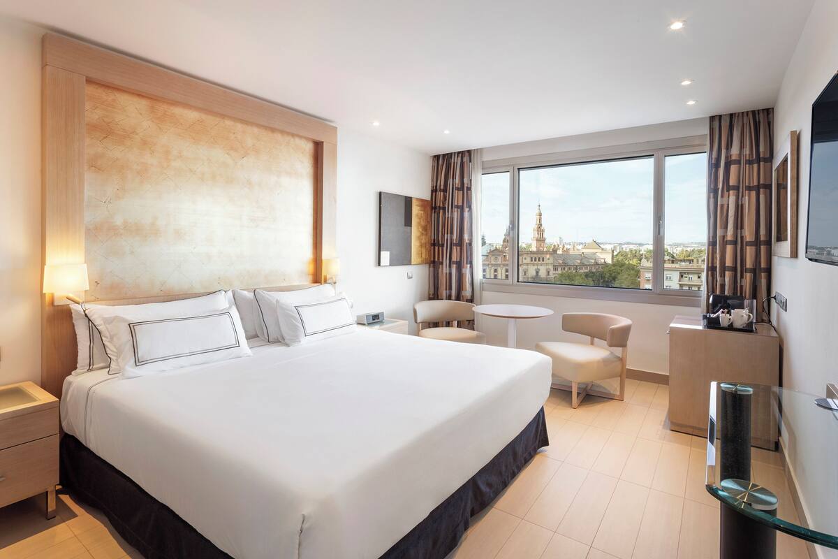 A hotel room with a large bed, a desk, a chair, and a view of a cityscape through the window.