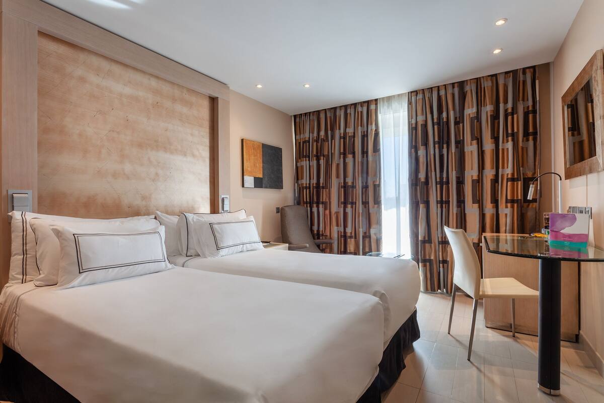 A hotel room with two beds, a desk with a chair, a large window with curtains, and a painting on the wall.