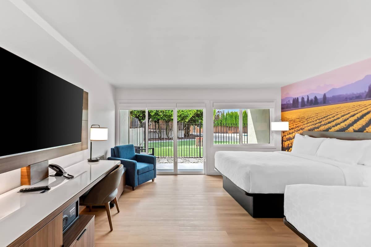 A hotel room with a bed, a desk with a chair, a TV, and a large window with a view of a field.