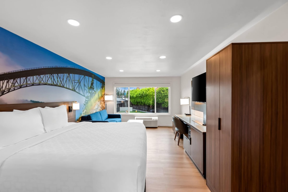 A modern hotel room with a large bed, a desk, a TV, and a mural of a bridge.