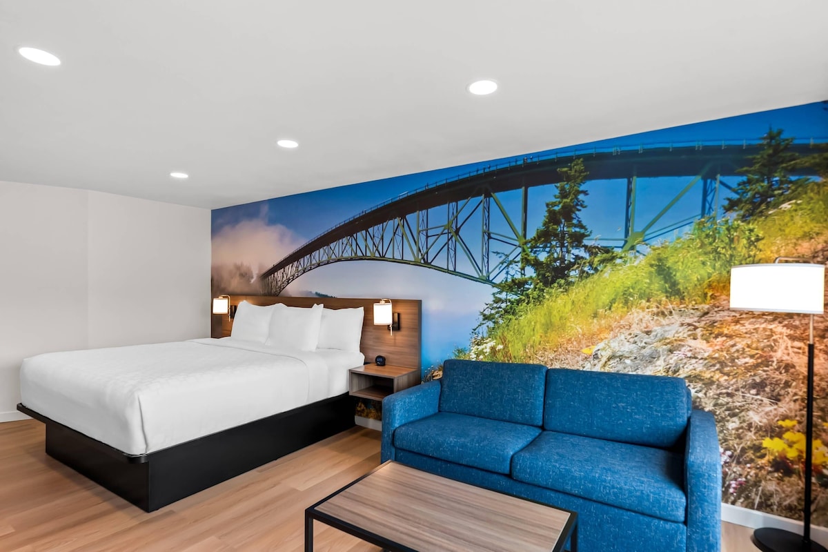 A hotel room with a bed, a blue sofa, a wooden coffee table, and a mural of a bridge.